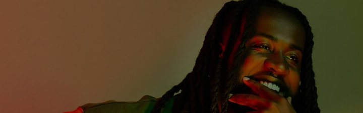 D'Juan Brundidge sitting infront of a multicolor background smiling with his hand to his mouth.